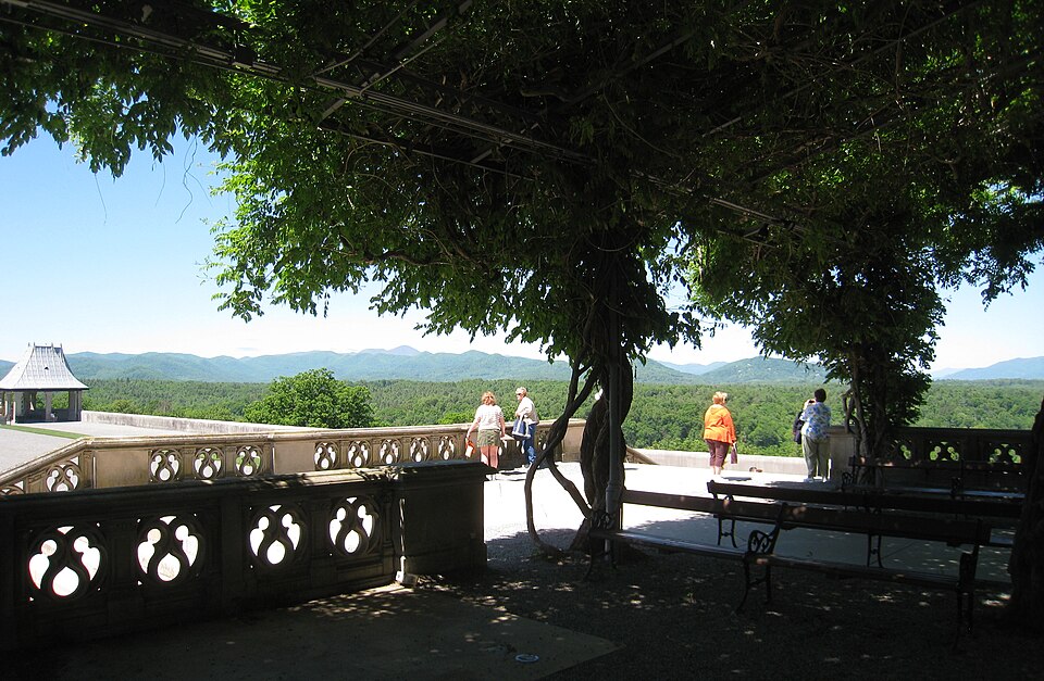 Visitors on the South Terrace at Biltmore House, under a wisteria-covered pergola, looking out over forested ridges toward the Blue Ridge Mountains