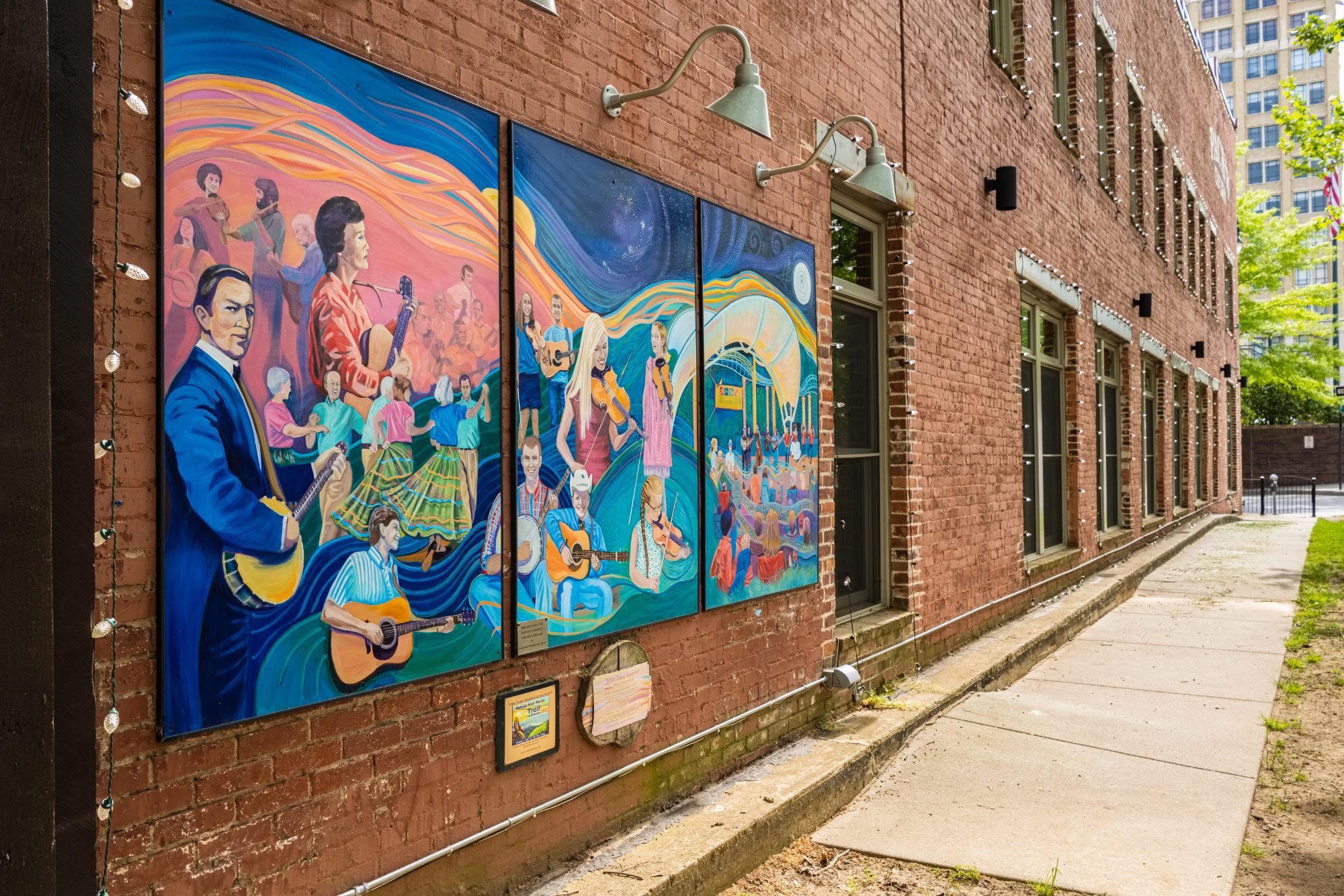 Three tall painted mural panels mounted on a red-brick warehouse wall showing a banjo player in a blue suit, a woman with a guitar, dancers in full period skirts mid-step and further banjo, guitar and fiddle players, all carried on broad swirling ribbons of orange, blue and green, with gooseneck lamps above and the sidewalk receding to a modern high-rise