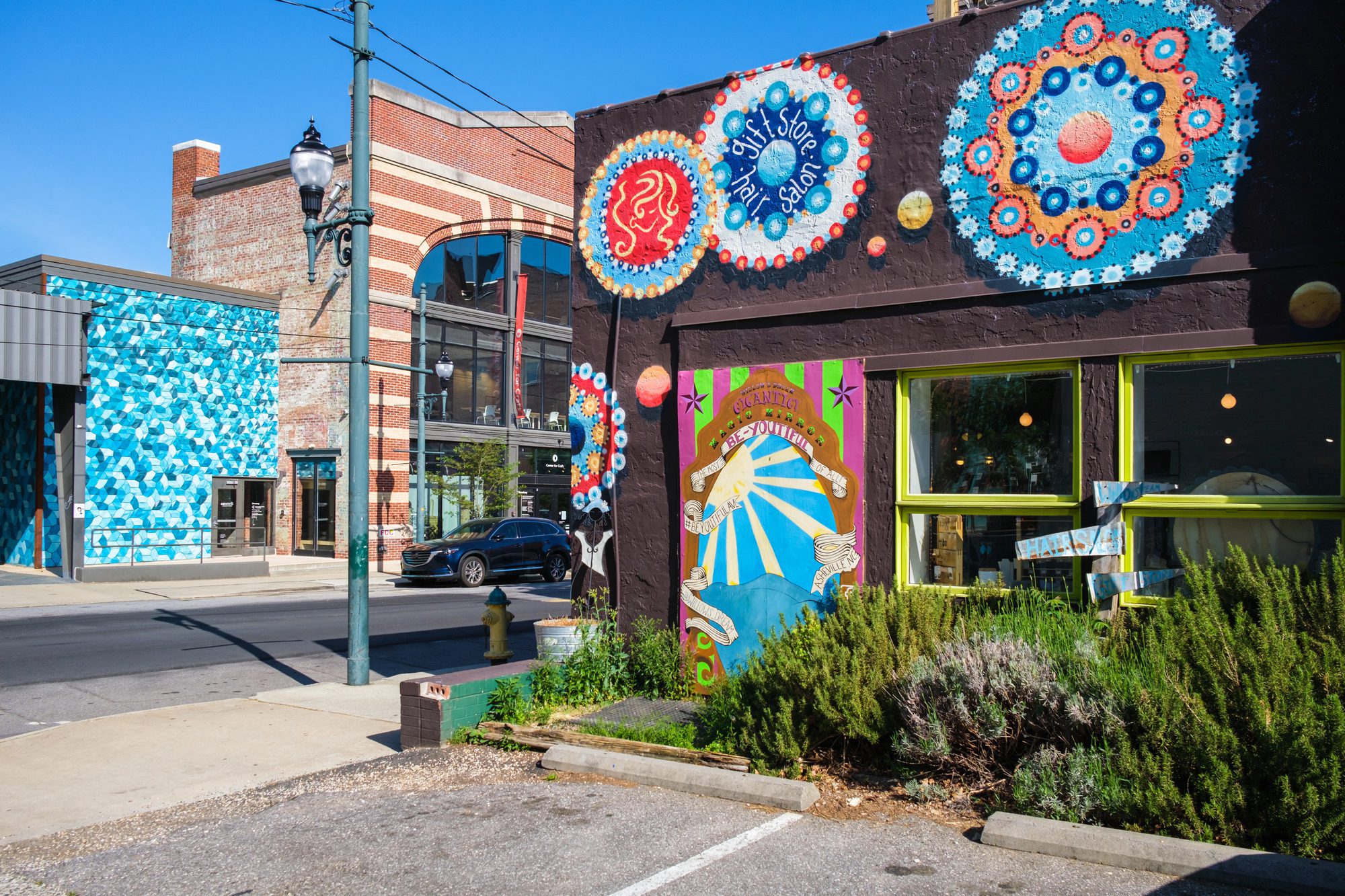 A dark aubergine-painted brick storefront with chartreuse-green window frames, five large circular mosaic and tile medallions in cobalt, white, orange and red fixed to its upper wall and parapet, rosemary and lavender in a raised bed in front, and a bright turquoise geometric mural on a wall across the street