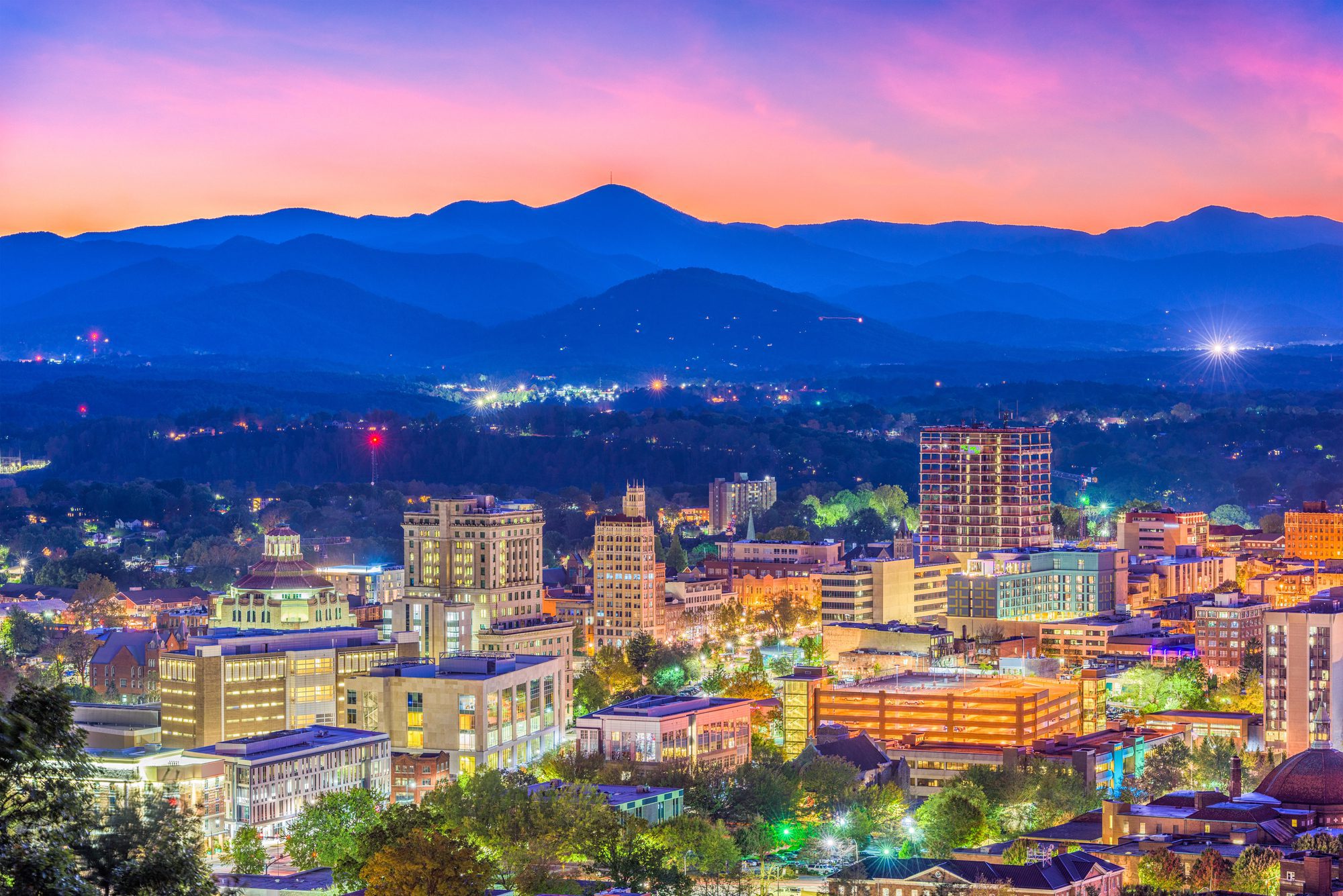 Downtown at blue hour photographed from above, lit office towers and brick blocks glowing across the lower two-thirds with street light and tree canopy threaded between them, three or four distinct blue mountain ridges rising behind with one prominent rounded peak, under a broad sunset gradient from deep blue through violet to hot pink