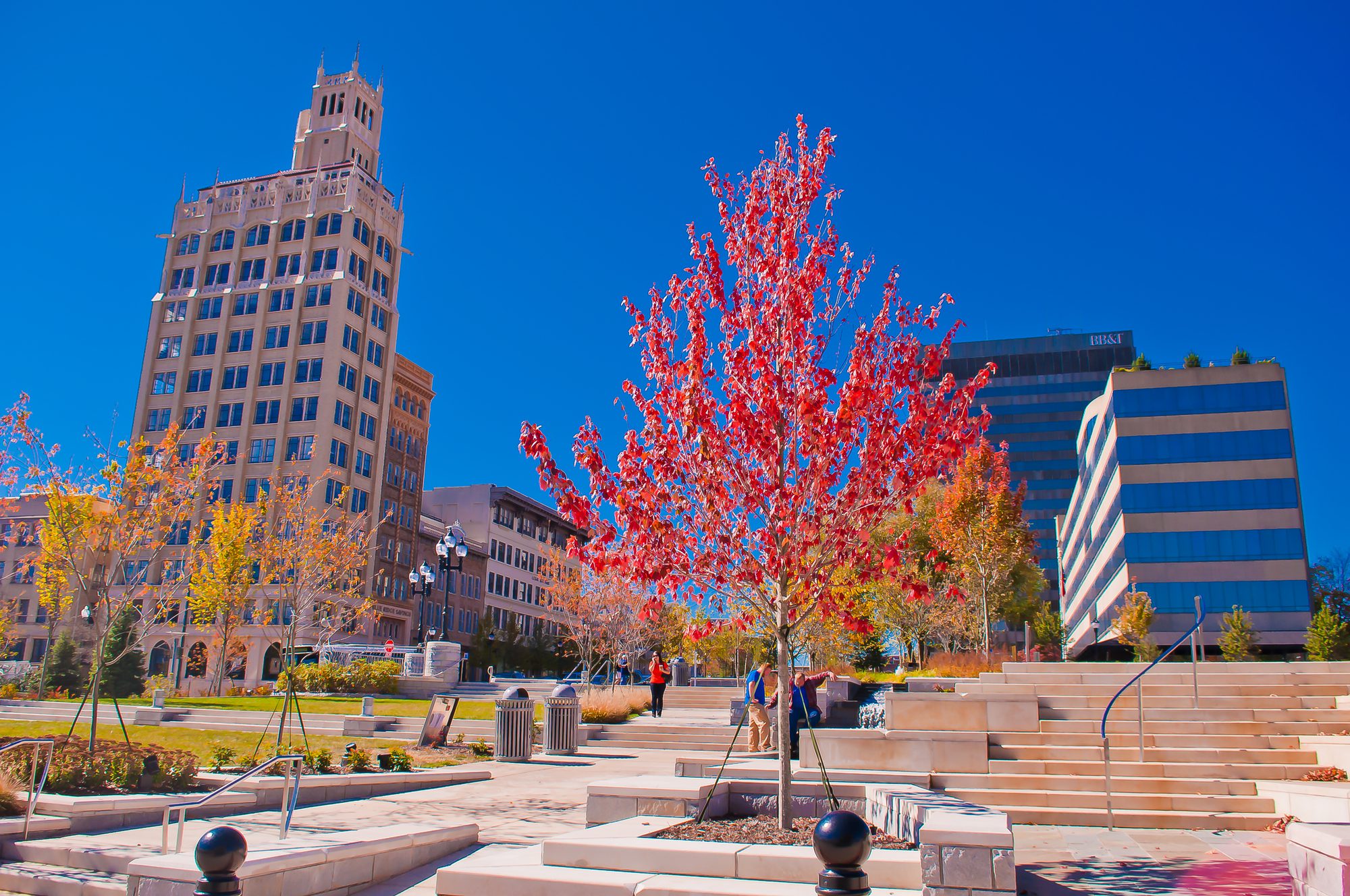 A narrow buff-stone high-rise rising through a corbelled parapet to an open belfry stage with crocketed pinnacles at each corner, a maple in brilliant scarlet leaf in front of it, a modern office block banded in pale stone and blue glass to the right, a stepped public plaza below