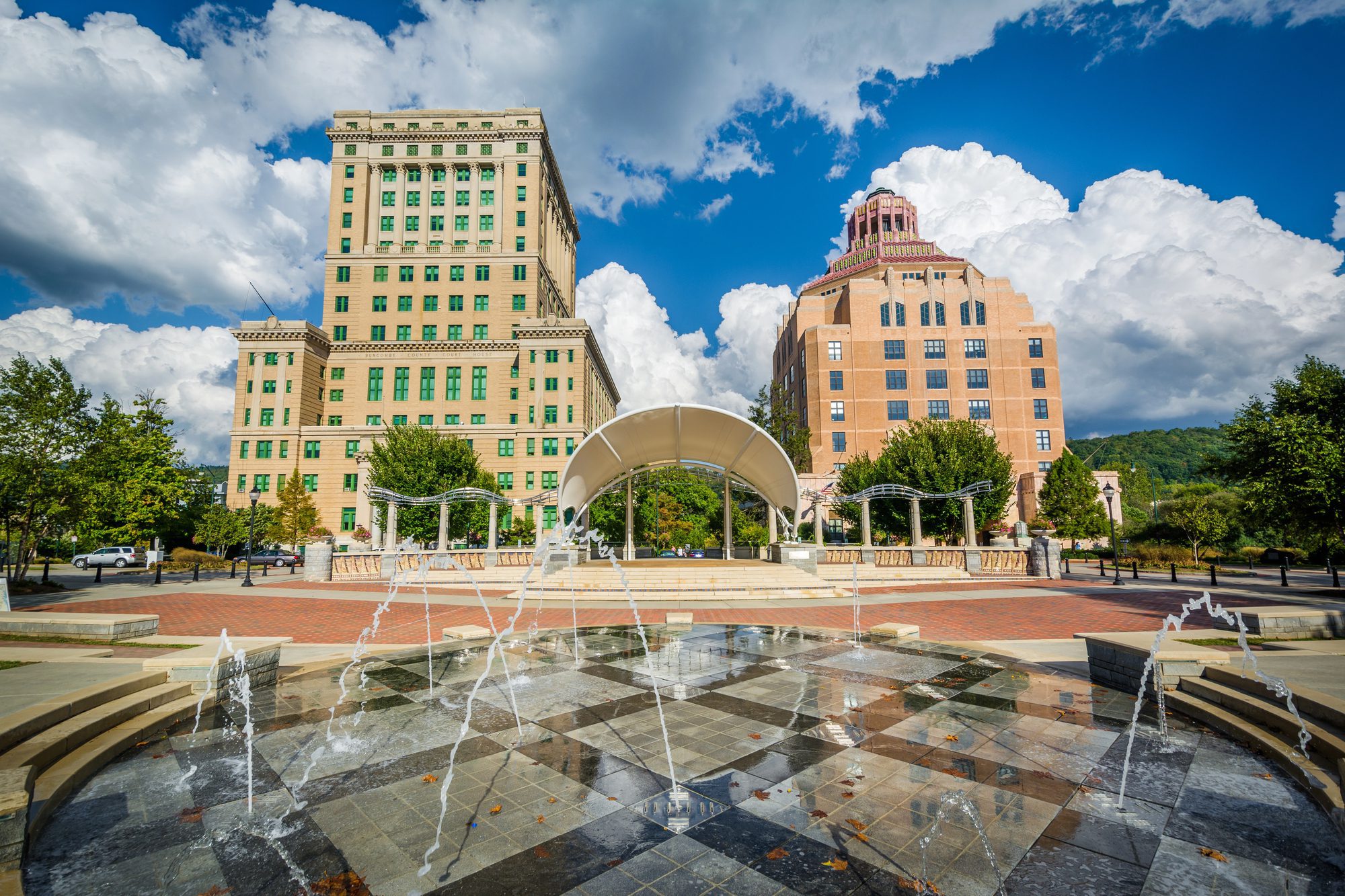 A checkerboard granite fountain basin throwing a dozen low jets with autumn leaves floating on the water, a white fabric-sail bandshell and classical colonnade behind, and two towers beyond - a buff Neoclassical high-rise with green-framed windows and a broad brick Art Deco block crowned with a stepped octagonal roof of pink and green tile