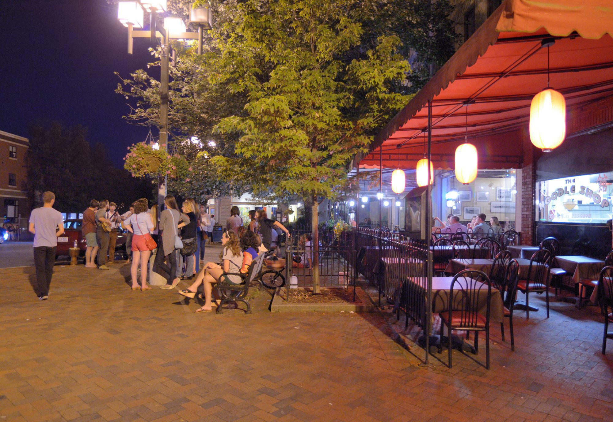 Dusk on a brick-paved sidewalk with twenty or more people standing, queueing and sitting on a bench under a large leafy tree lit from below, beside an outdoor restaurant terrace under a deep orange awning hung with four glowing globe pendant lanterns, its iron chairs empty in the foreground and the lit interior full of diners