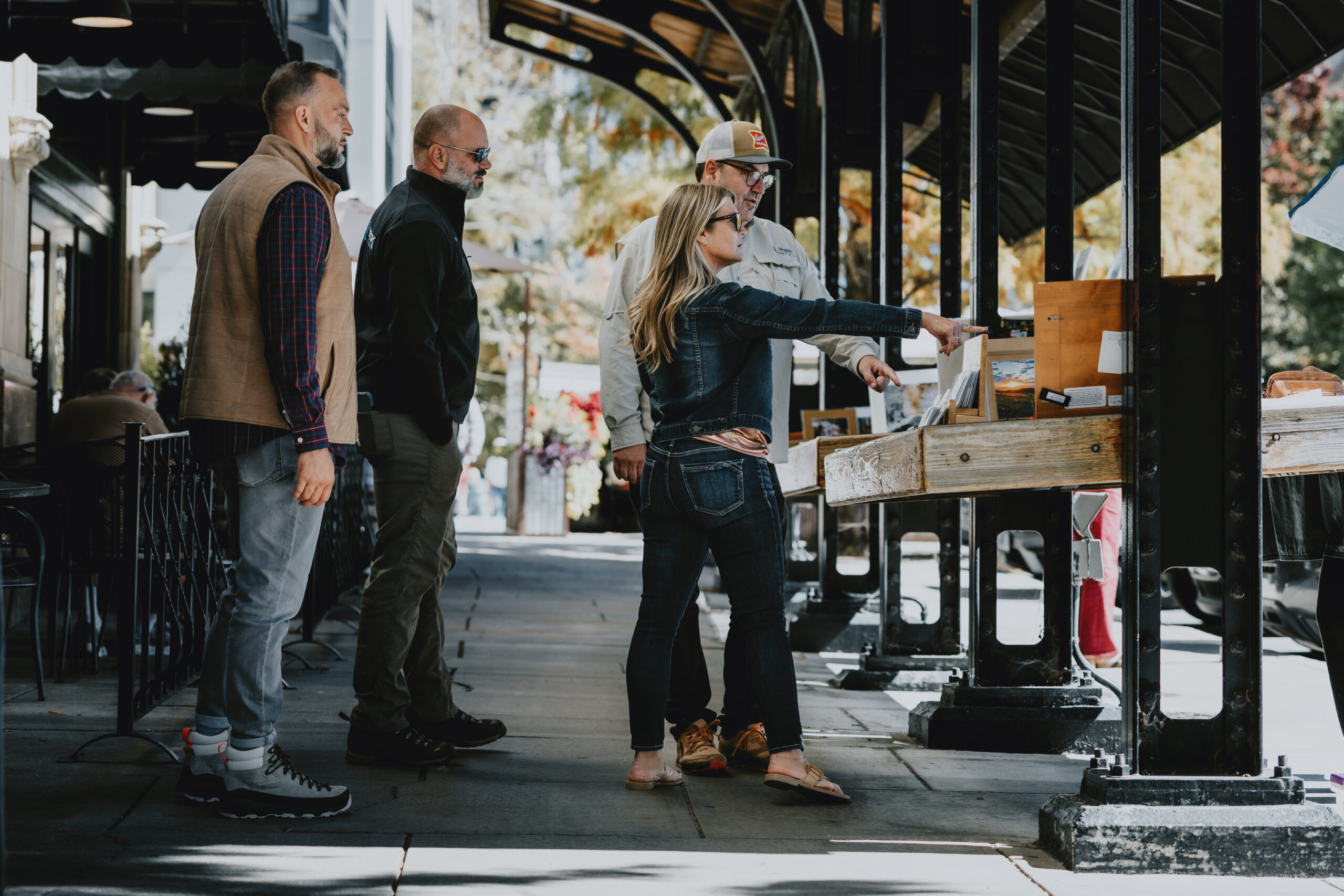 A guide and four guests on a downtown pavement under a black cast-iron and glass canopy, one guest pointing at framed prints propped on a timber vendor's stall, cafe railings and autumn foliage behind