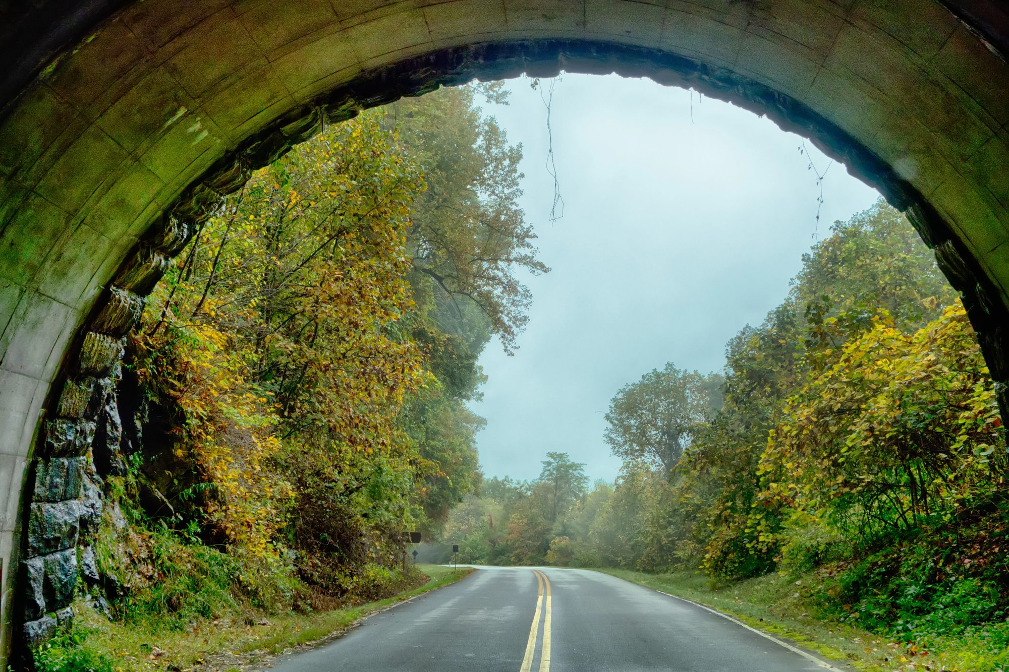 The view out through a stone tunnel arch to wet road and autumn trees