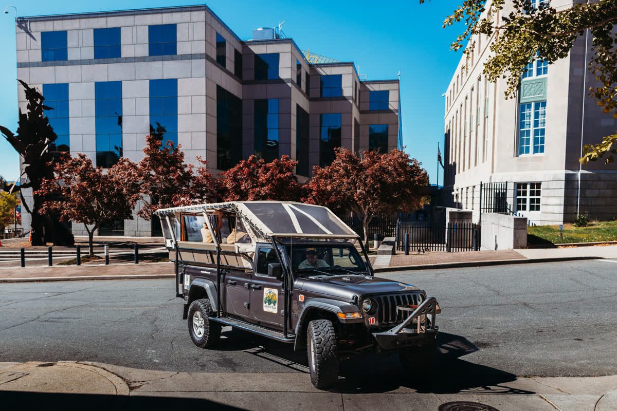 The open-air Jeep Gladiator in three-quarter view on a downtown street with the guide at the wheel, a modern granite-and-blue-glass office block behind it and a pale limestone building with tall green-framed windows to the right, Japanese maples in deep red autumn leaf
