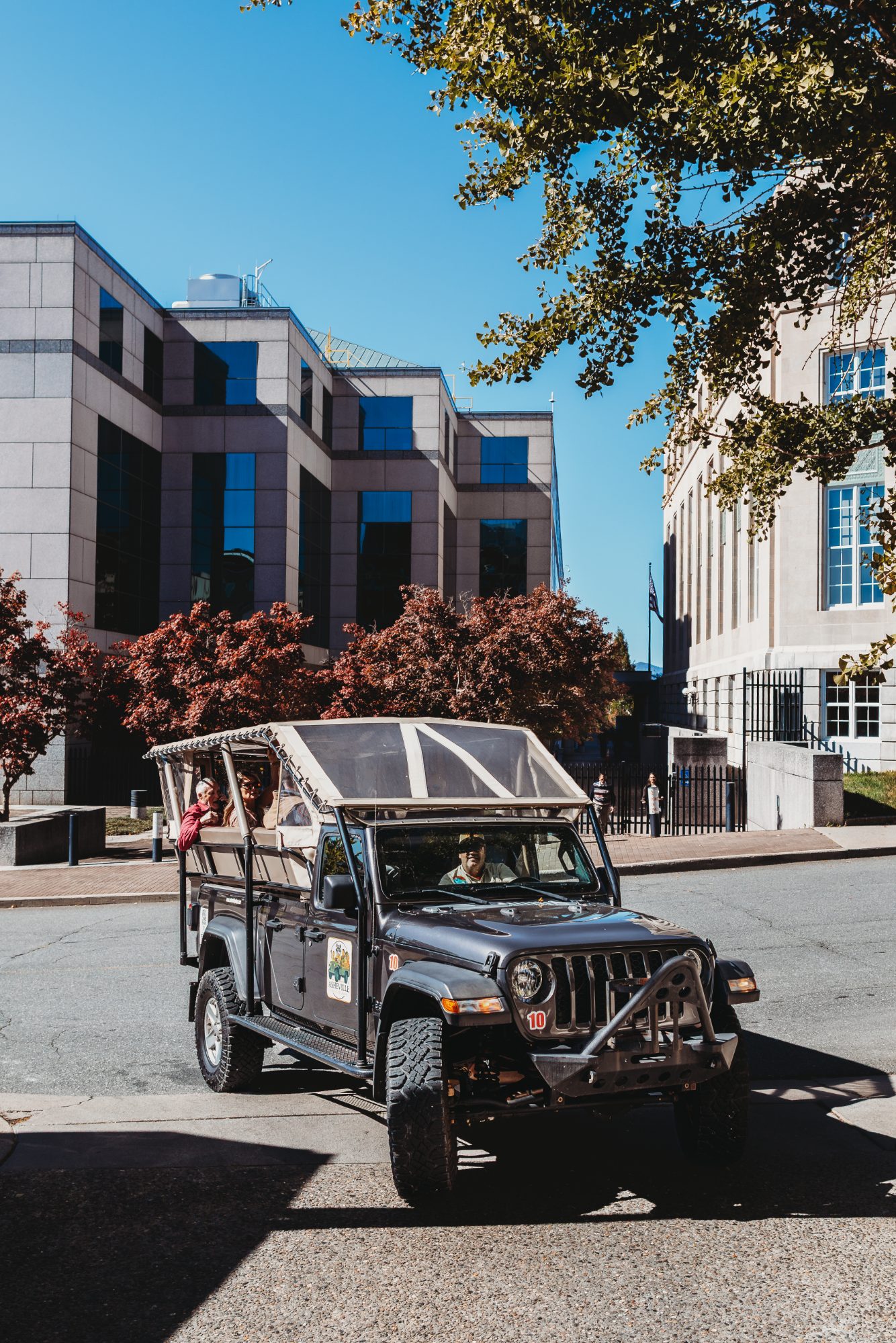The same open-air Jeep Gladiator photographed upright from lower down, two guests clearly seated on the rear bench with the guide at the wheel, the granite-and-blue-glass block filling the upper frame and red Japanese maples across the middle