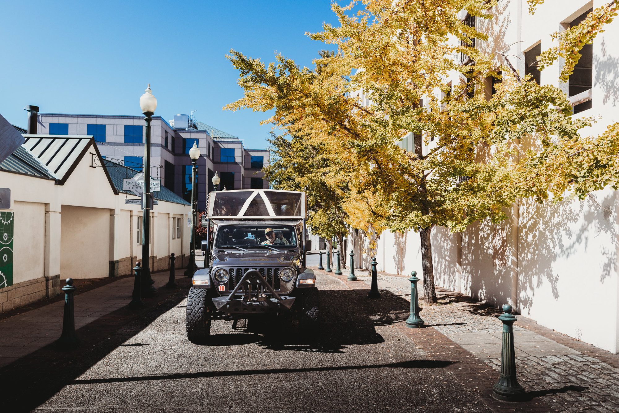 The operator's dark grey Jeep Wrangler with a cream soft top parked on a brick-paved downtown pedestrian lane between cast-iron bollards and street lamps, a row of ginkgo trees in full autumn gold along the right-hand wall