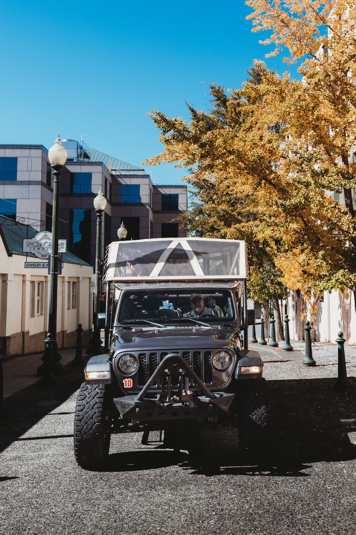 The open-air Jeep Gladiator front-on and close on a brick lane, the driver visible through the windscreen, a gold ginkgo canopy filling the upper right and an ornate twin-globe street lamp standing centre-left