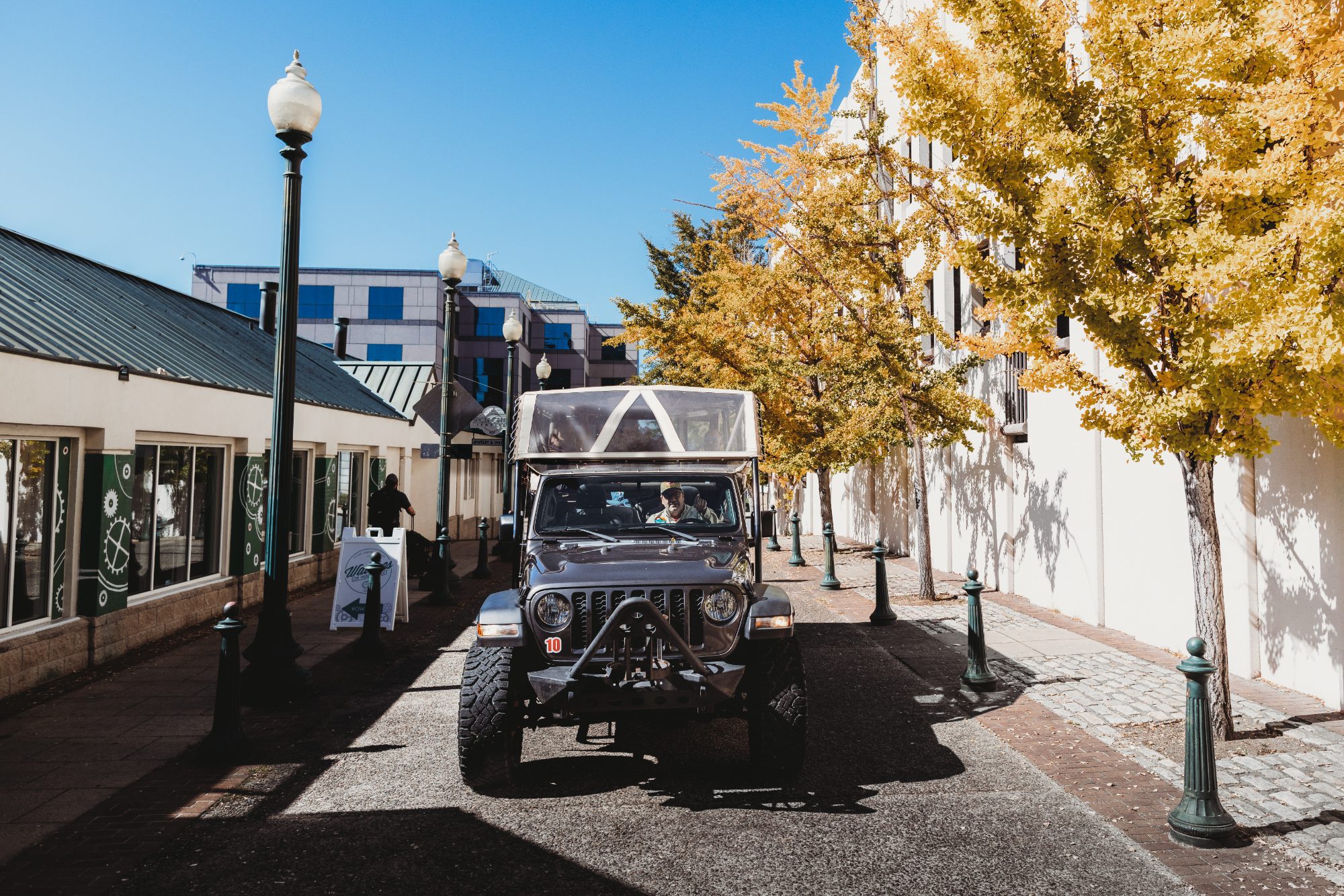 The operator's open-air Jeep Gladiator head-on in a brick-paved pedestrian lane, a guest in the passenger seat, an enormous ginkgo in full gold leaf to the right, ornate twin-globe street lamps and green cast-iron bollards down both sides