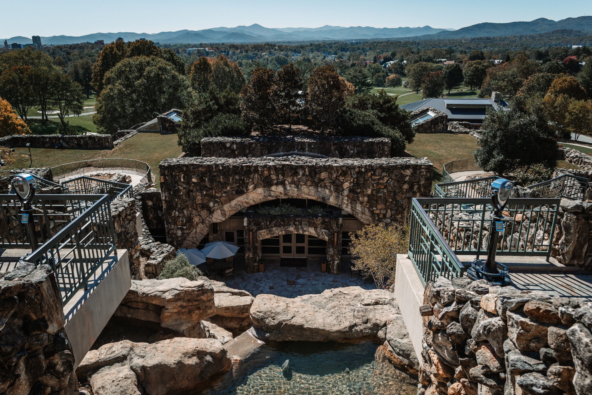 An elevated stone terrace with green metal railings and two coin-operated viewing binoculars above a massive stone arch, looking out over treetops in early autumn colour, a distant city skyline of low towers, and layered blue mountain ridges across the horizon
