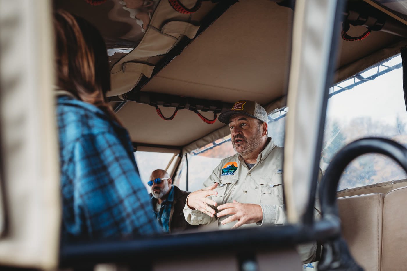 The guide photographed from a passenger seat, hands raised open mid-explanation and speaking, a guest in a blue check shirt in the foreground and a second guest in sunglasses listening behind him, tan canopy and roll cage overhead