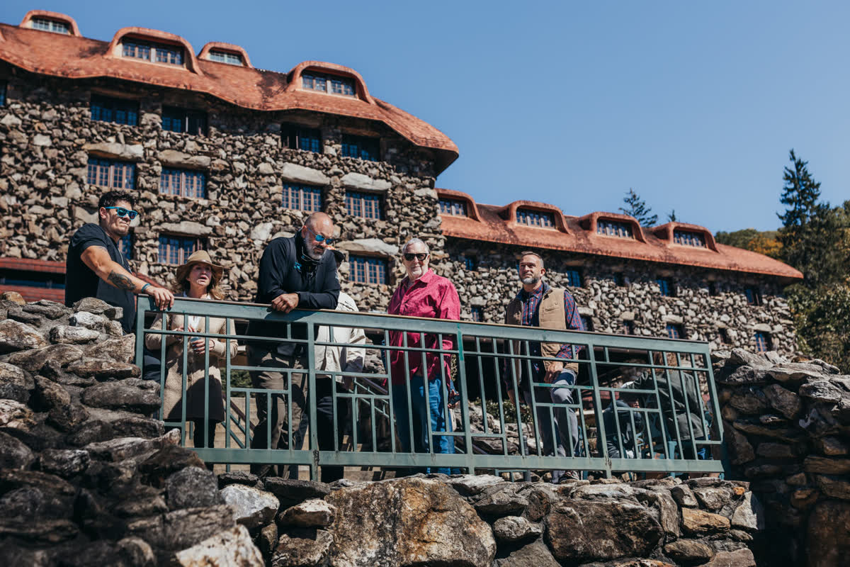 Five guests and their guide leaning on a green metal railing on a stone terrace built into a wall of massive boulders, a large rough-stone building with a deep red tiled roof and rows of dormer windows rising behind them
