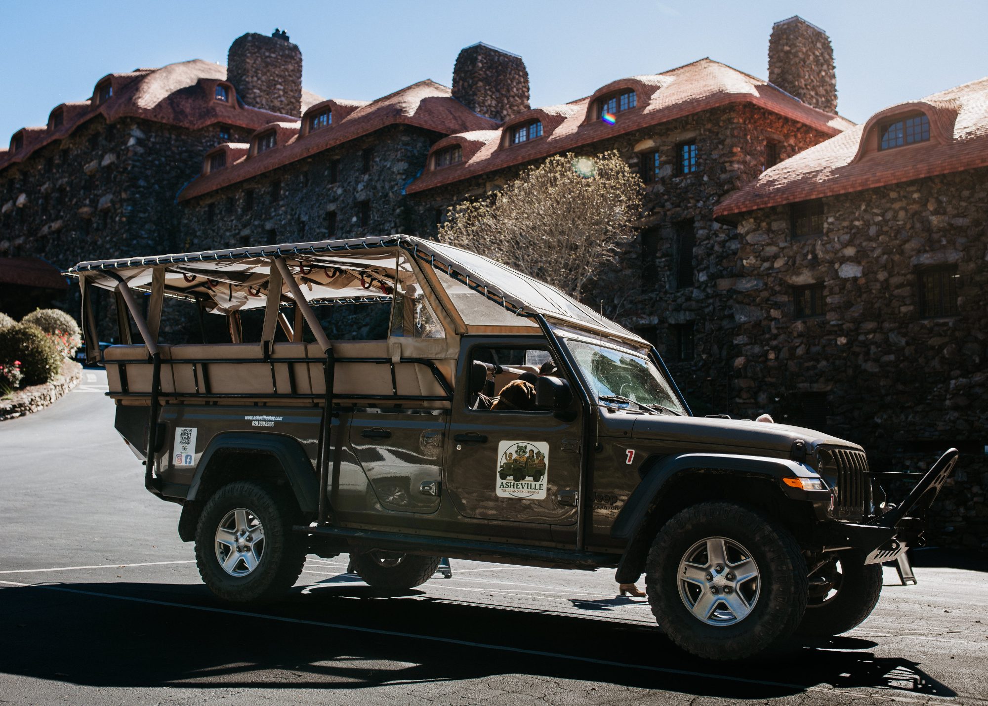 The operator's dark olive open-air Jeep with a tan canvas canopy and two rows of bench seating, parked in front of a large stone building with a red tiled roof and stone chimneys under a clear blue sky