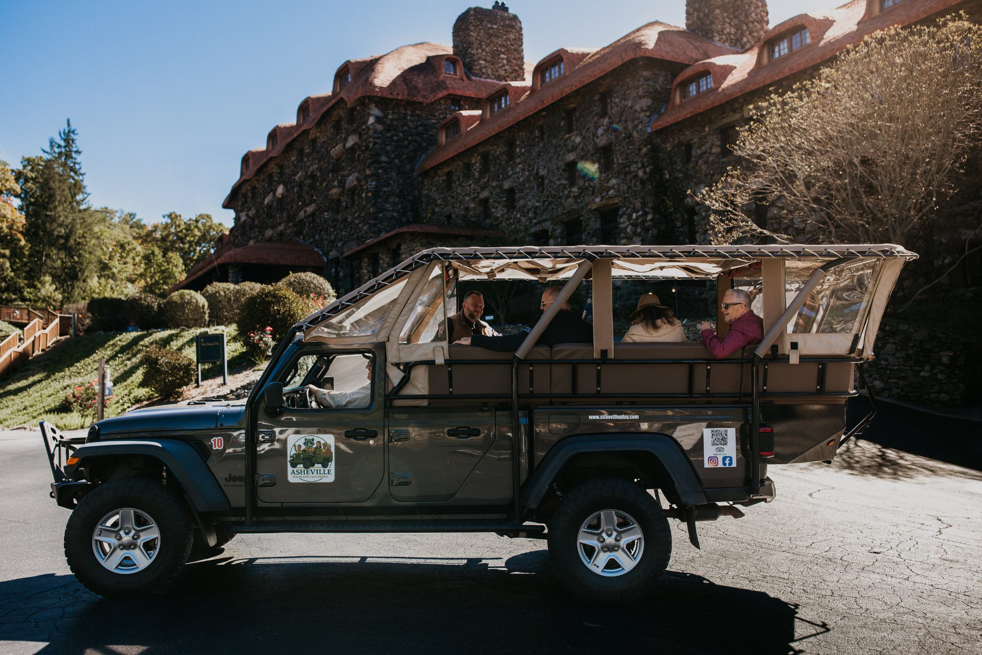 The operator's dark olive open-air Jeep Gladiator in profile with a tan canvas canopy and clear vinyl side panels rolled open, four guests seated on two bench rows behind the cab, the company logo and web address on the door