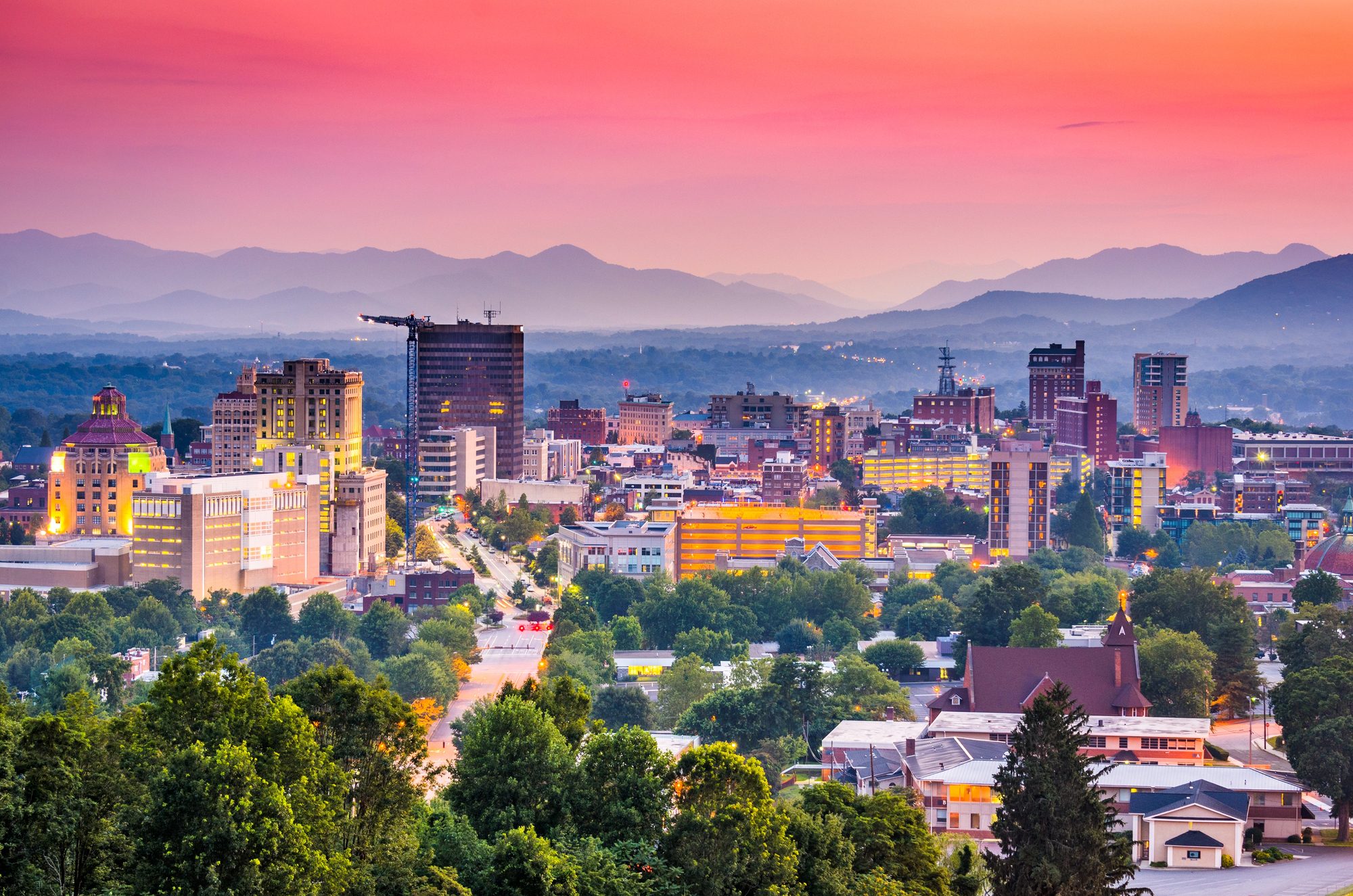 A mountain city skyline at dusk under a pink and orange sky, long ridgelines behind it
