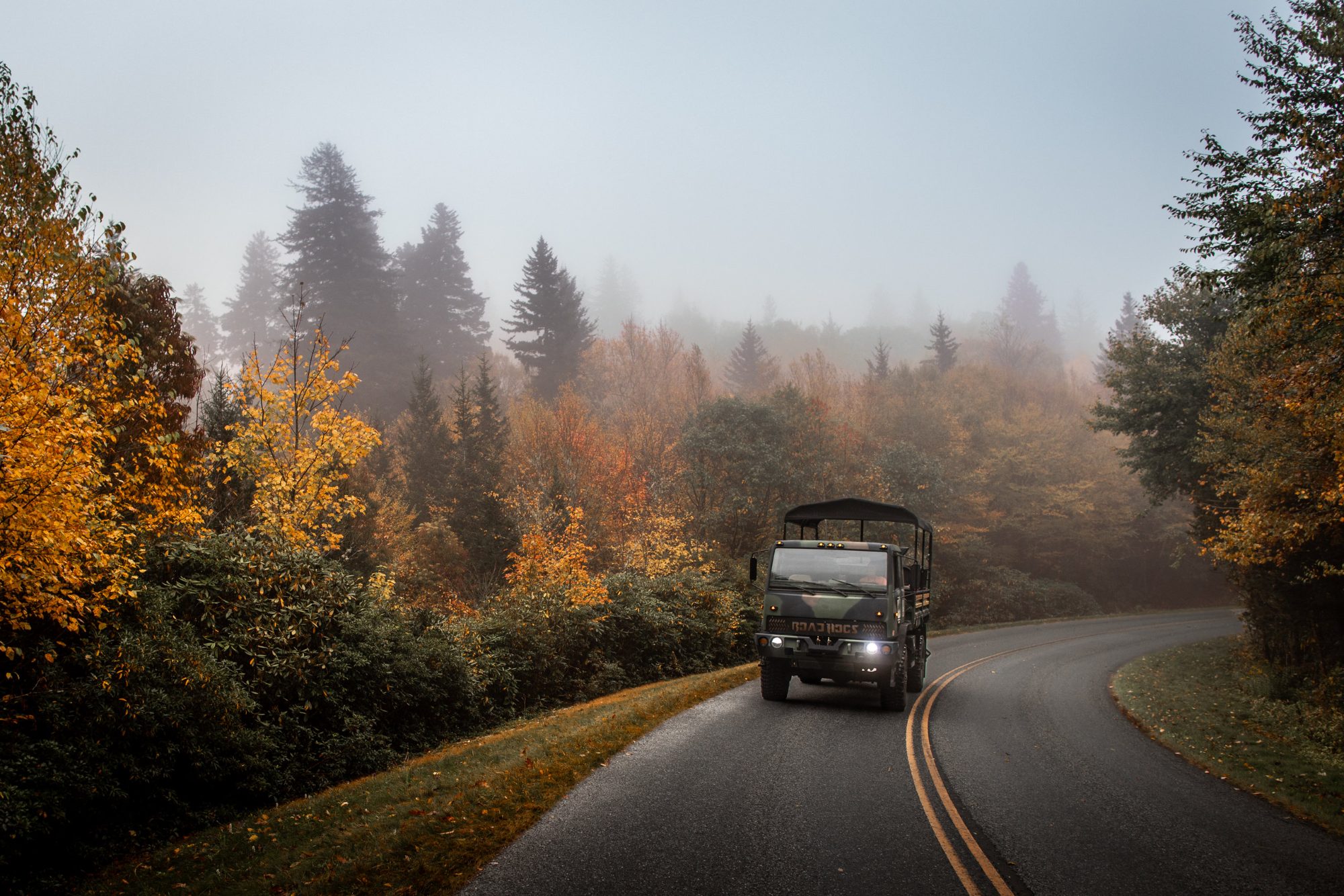 The operator's large open-air troop-carrier tour vehicle on a two-lane mountain road in autumn fog, headlights on