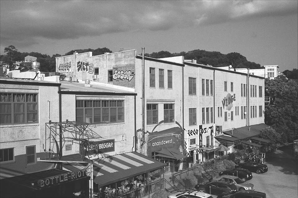 A black-and-white view across River Arts District rooftops: brick industrial buildings with graffiti, painted signs reading Wedge and Bottle Riot, awnings and parked cars along the street below a wooded hill