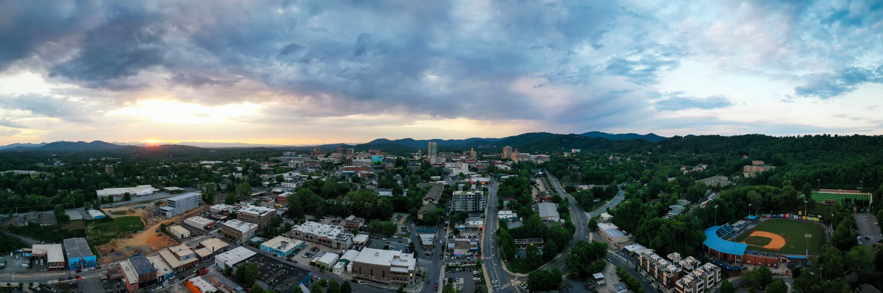 Downtown Asheville from the air at sunset: a compact cluster of towers in a bowl of green canopy, a baseball park at the right edge, roads threading through trees, and layered blue mountain ridges running the whole horizon under a breaking sky