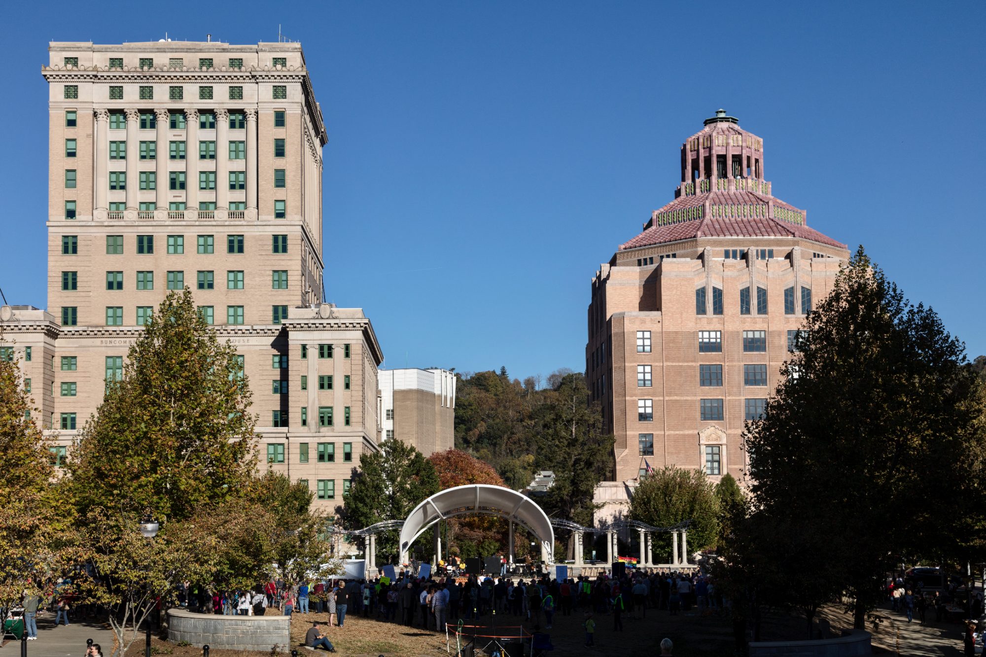 Two civic towers over a downtown park: a pale limestone Neoclassical high-rise with an engaged colonnade whose frieze reads Buncombe County Courthouse, and a brick Art Deco tower crowned with a stepped octagonal roof of pink and green tile, a crowd gathered at a bandshell below