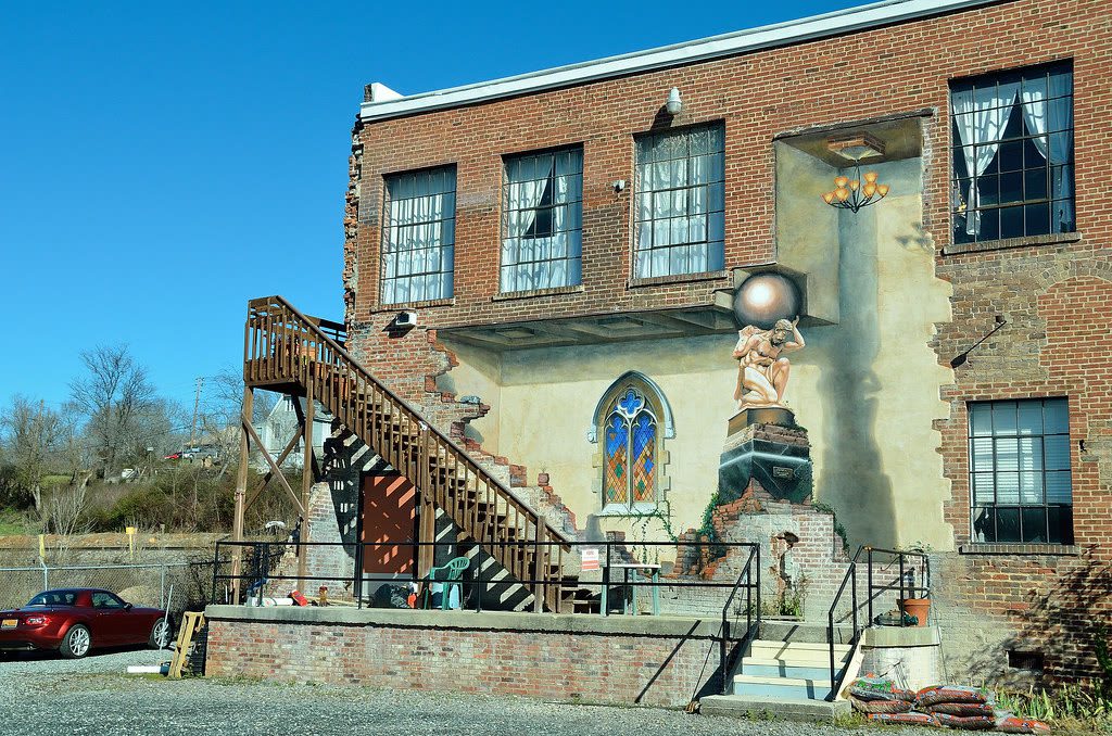 Cotton Mill Studios in the River Arts District: a two-storey brick mill building with a wooden exterior staircase and a trompe-l'oeil mural of a figure holding a sphere beside a painted stained-glass window