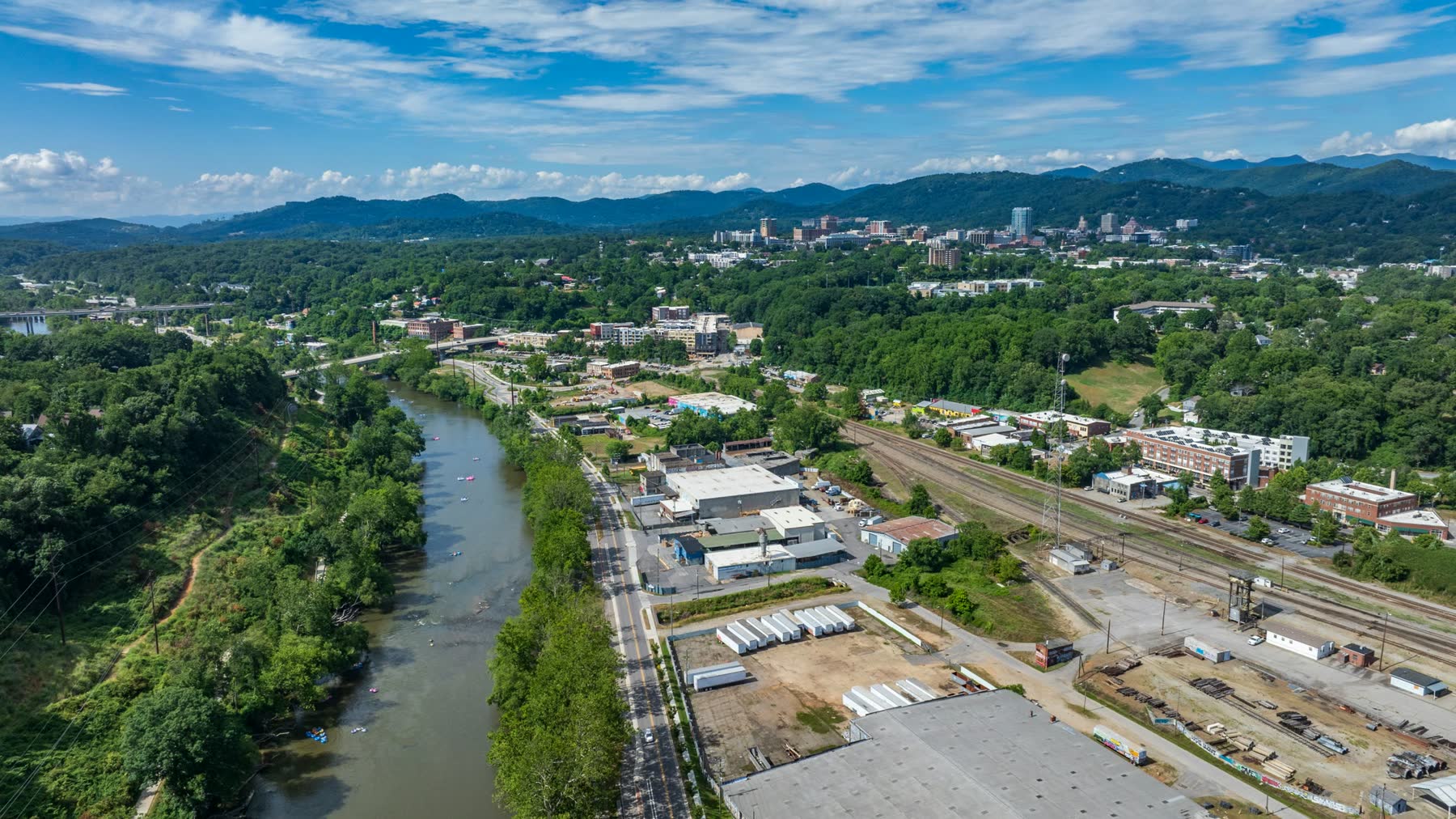 An aerial summer view of the French Broad River threading past the River Arts District, tubers floating as small bright dots on the water, downtown Asheville and layered ridges in the distance