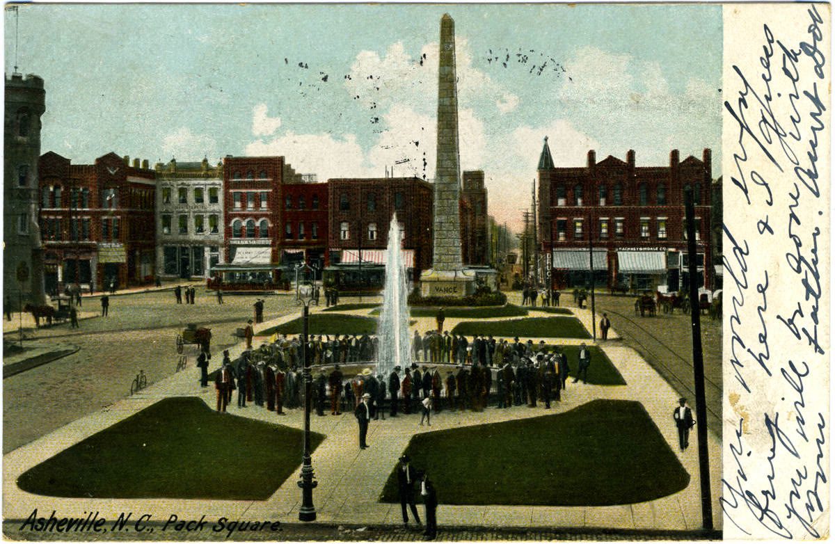A hand-tinted early-1900s postcard printed Asheville, N.C., Pack Square: a crowd in hats around a tall fountain jet, the granite Vance obelisk behind, brick storefronts and streetcars ringing the square, handwriting in the margin