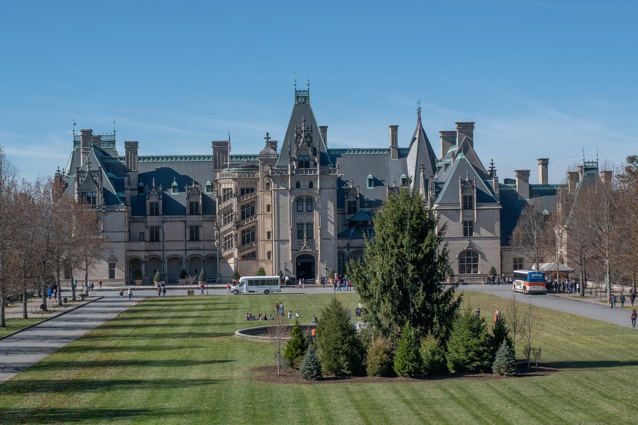 The Biltmore House facade across its front lawn: a French Renaissance chateau of pale stone with steep slate roofs, ornate dormers and chimneys, tour shuttles and visitors small at its entrance
