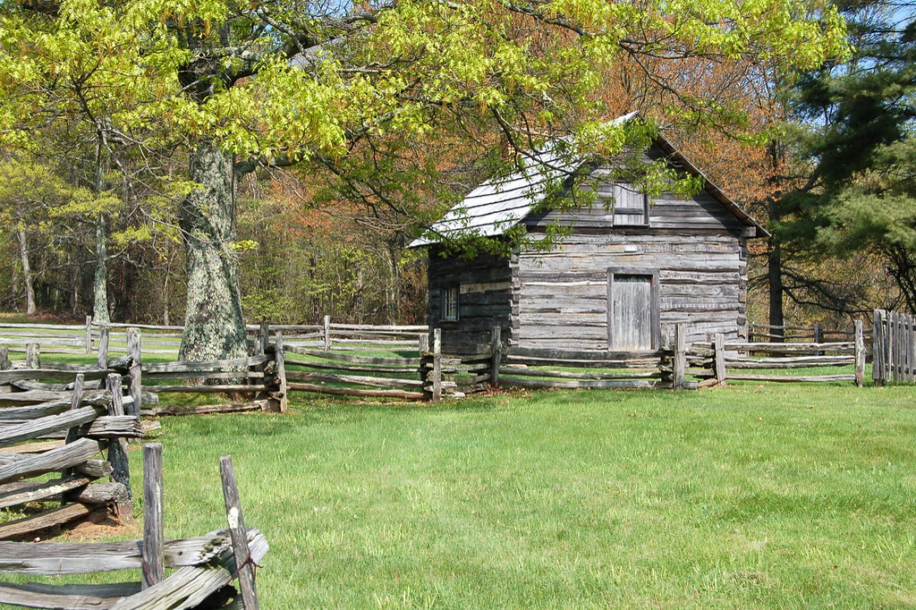 Puckett Cabin, VA. A very small chinked-log one-room cabin with a stone chimney standing alone in a mown clearing at the roadside.