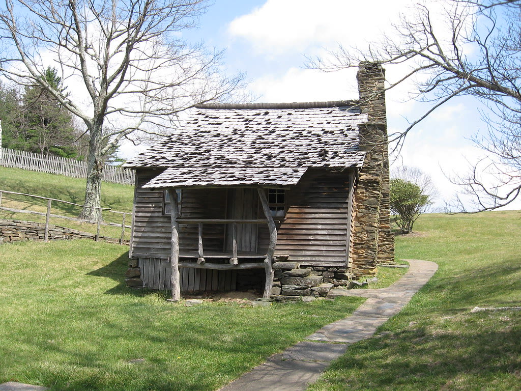 Brinegar Cabin, NC. A small dark-log cabin with stone chimney on an open ridgetop meadow, mountains falling away behind.