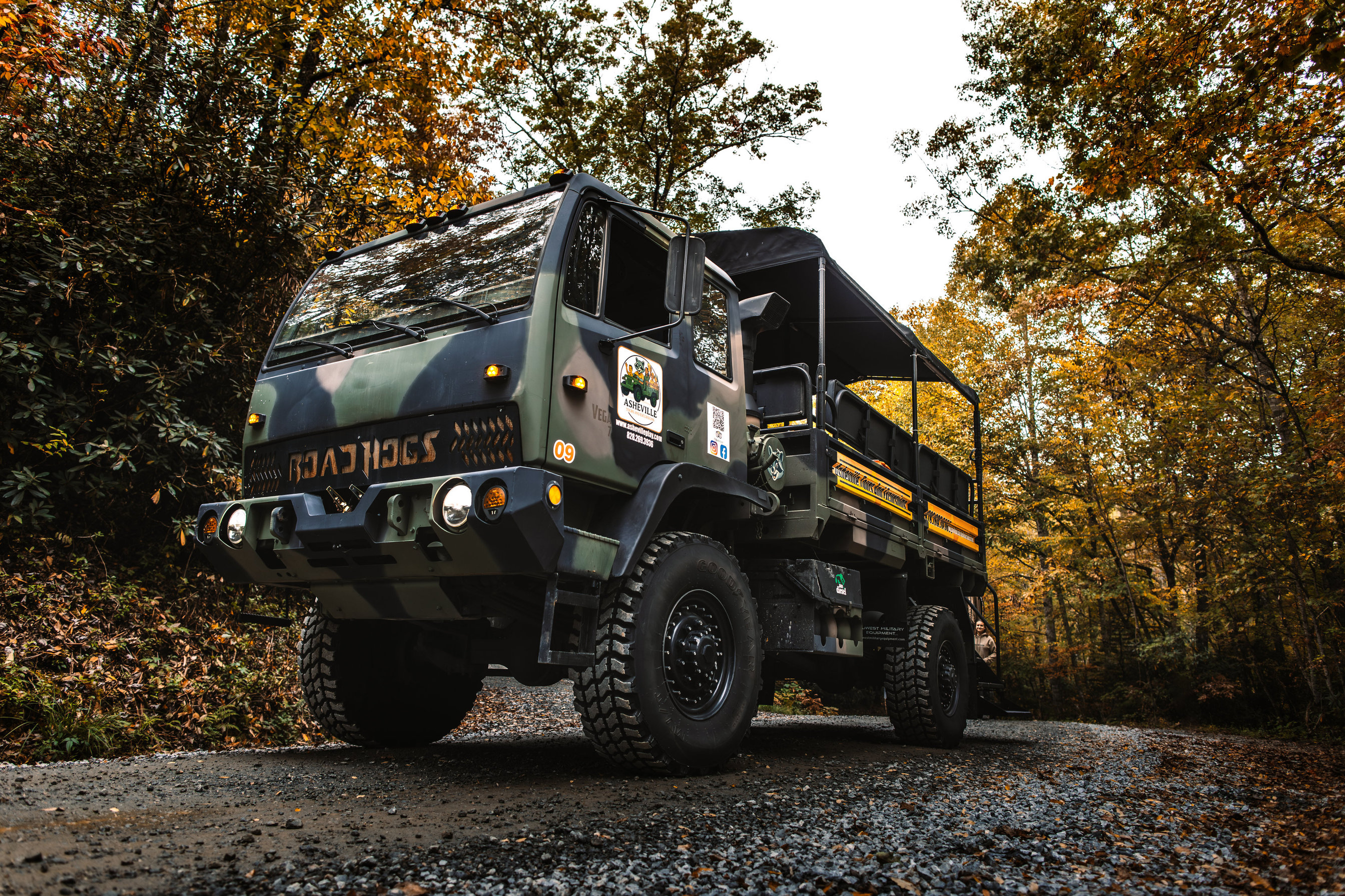 A camouflage open-air troop-carrier truck with bench seating under a canopy frame, photographed low from the front three-quarter
