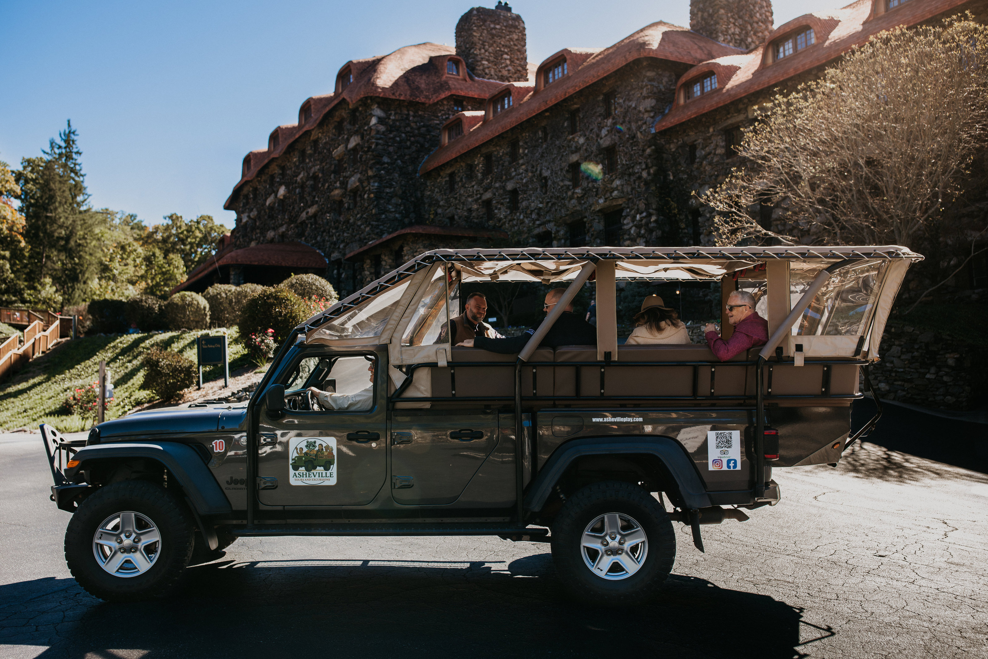 An open-air Jeep Gladiator in profile on a sunlit autumn forest road, four guests seated on bench seating in the open bed beneath a canvas and clear-vinyl canopy, operator branding on the door