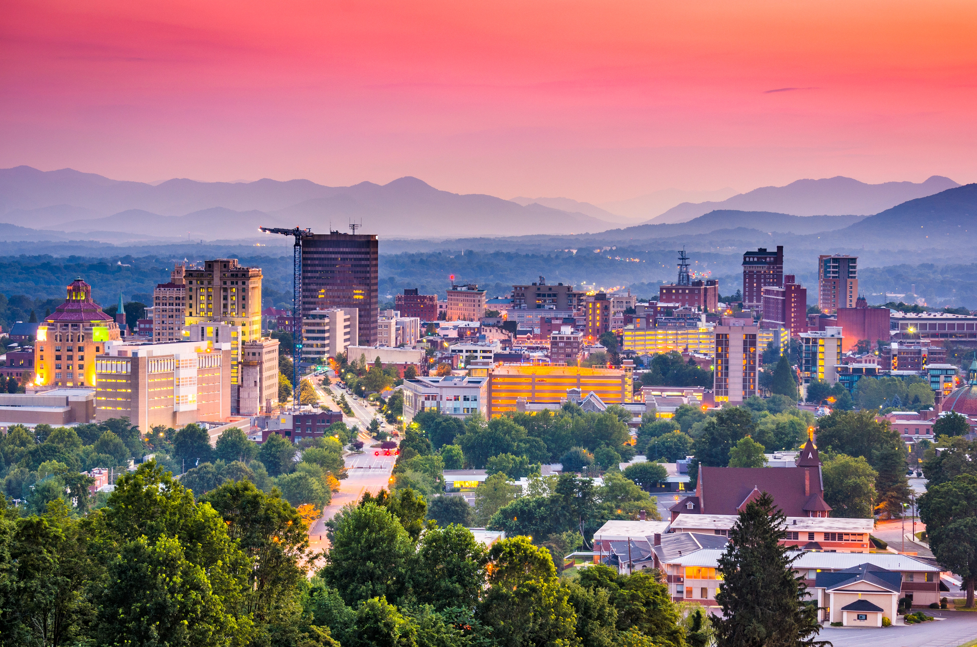 A mountain city skyline at dusk under a pink and orange sky, long ridgelines behind it
